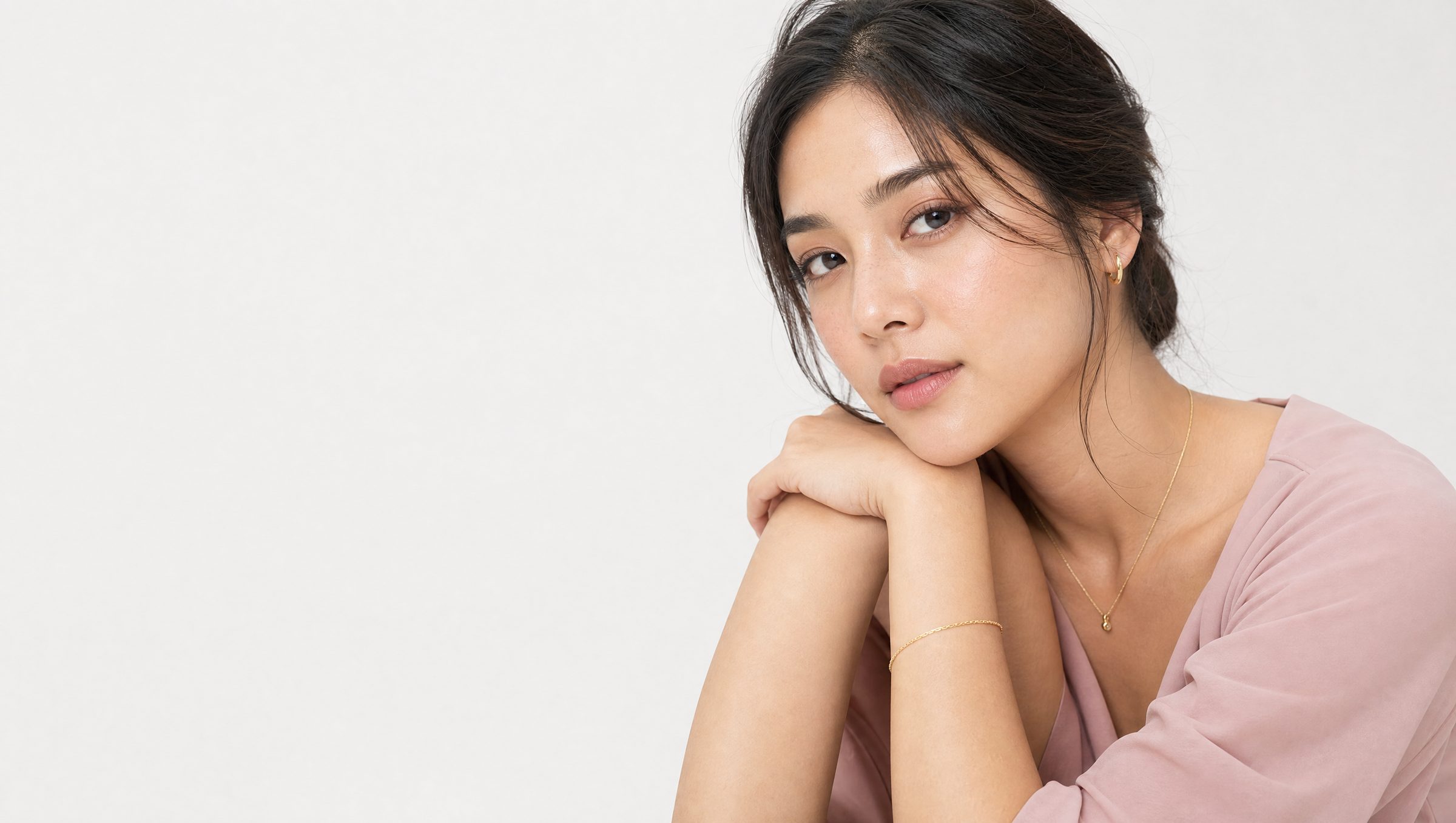 Portrait of a woman resting her chin on her clasped hands against a plain light backdrop, wearing a small gold hoop earring, a delicate gold necklace with a teardrop pendant, and a thin gold bracelet.